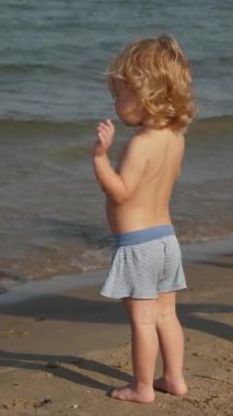 Kumlu kıyı şeridini keşfeden çocuk, kumsalda oynayan küçük sarışın çocuk, deniz kenarında eğlenen küçük sarışın çocuk, yaz sıcağını yaşayan genç kaşif, kıyı şeridinde gezinirken ve su sıçratırken.
