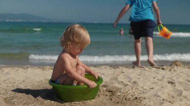 Küçük çocuk güneşli tropikal sahilde küvette oturuyor, sevimli bir çocuk kumsalda kumun ve suyun tadını çıkarırken, biri yaz günü elinde kovayla ağır çekimde koşuyor.
