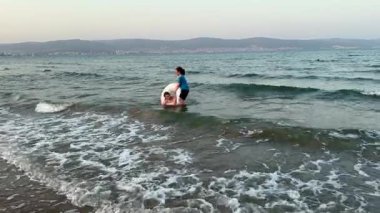 Sahil şeridinde oynayan genç ve çocuk, hafif dalgalar ayak bilekleri, ılık günbatımı ışığı, köpükle çizilmiş sahil, yaz tatili havası, samimi aile bağları