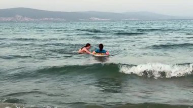 Ergen ve çocuk eğleniyor. Yaşlı olan, yumuşak kıyı sularında çocuğa yardım ediyor. Bakıcı, şişme yüzükle sakin denizde çocuğa rehberlik ediyor. Kaygısız bir yaz akşamı, kardeş, çocuğun yumuşak sörf yapmasına yardım eder.