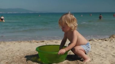 Neşeli sahil keşfi. Küçük bir çocuk kumsalda güneşli bir günün tadını çıkarıyor. Mutlu bir çocuk yaz eğlencesinde yeşil bir havuzda suyla oynar. Bebek mutlu bir şekilde güneşli, parlak bir sahilde suyla oynuyor.