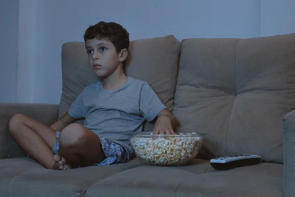 Menino pequeno no sofá assistindo TV e comendo pipocas à noite i — Fotografia de Stock