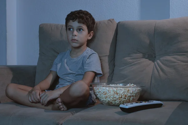 Niño pequeño en el sofá viendo la televisión y comiendo palomitas de maíz en la noche i — Foto de Stock