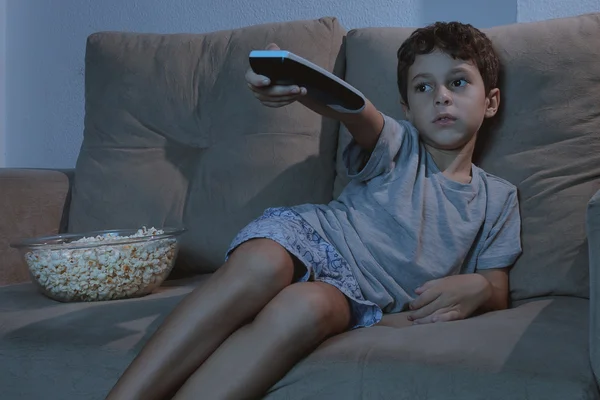 Anak kecil di sofa menonton TV dan makan popcorn di malam hari i — Stok Foto