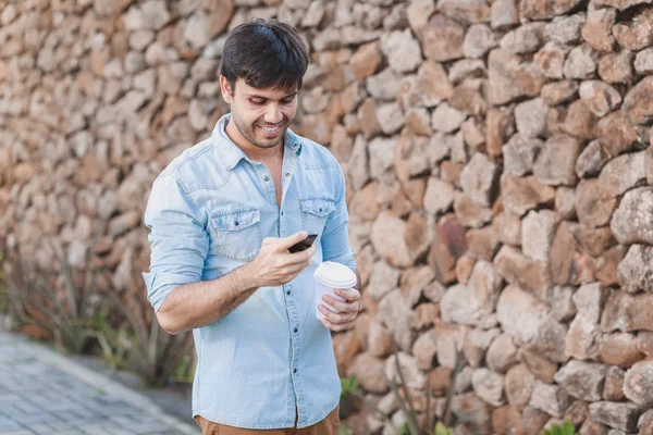 Портрет молодого человека, пьющего кофе на улице при помощи мобильного телефона — стоковое фото