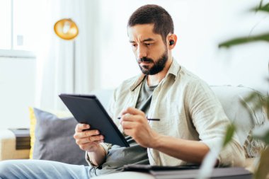 Evden çalışan sakallı genç bir adam. Evdeki koltukta dijital tablet kullanan bir adam.