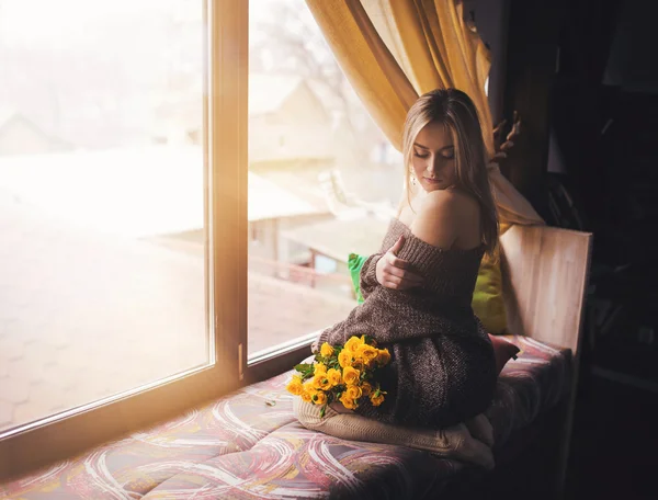 Retrato de jovem mulher bonita em pulôver cinza sentado na soleira da janela com flores amarelas — Fotografia de Stock
