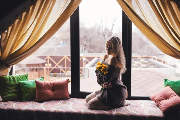 Portré, fiatal gyönyörű nő ül a sárga virágok az ablakpárkányon szürke pulóver — Stock Fotó