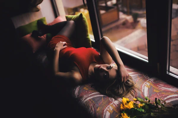 Retrato de la joven hermosa mujer en vestido rojo se encuentra en el alféizar de la ventana con flores amarillas — Foto de Stock