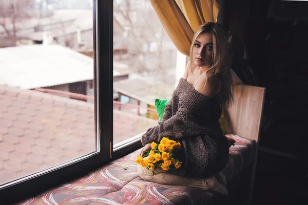 Porträt einer jungen schönen Frau in grauem Pullover, die auf der Fensterbank mit gelben Blumen sitzt — Stockfoto