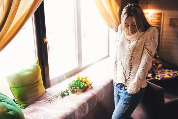 Hermosa joven sexy mujer con pelo largo y rubio con maquillaje natural usando suéter y jeans se encuentra cerca de la ventana — Foto de Stock