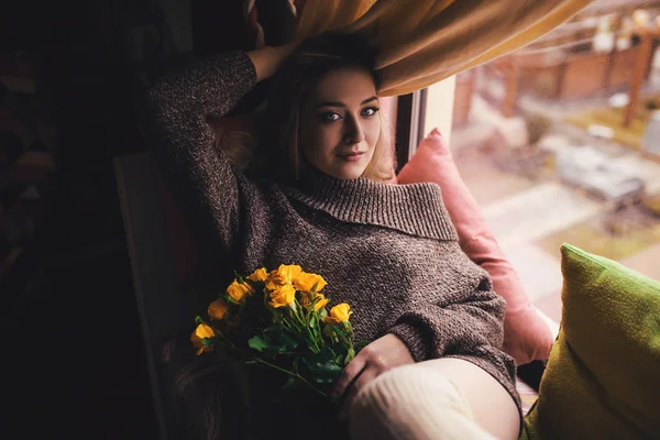 Potret wanita cantik muda dalam abu-abu pullover duduk di ambang jendela dengan bunga kuning — Stok Foto