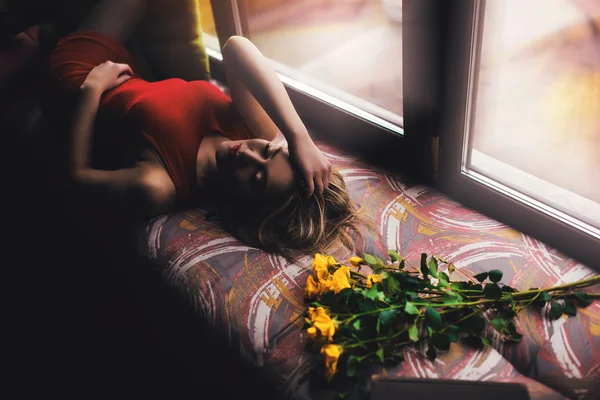 Porträt einer jungen schönen Frau in rotem Kleid liegt auf Fensterbank mit gelben Blumen — Stockfoto