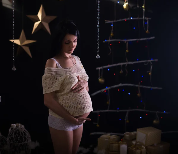 Retrato de hermosa morena embarazada sexy en peignoir transparente blanco, sosteniendo un sombrero para su bebé, el interior de año nuevo de la casa cerca del árbol de Navidad y la chimenea —  Fotos de Stock