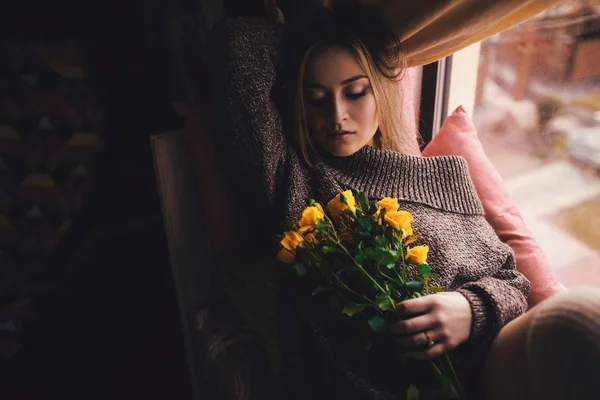 Retrato de mujer hermosa joven en jersey gris sentado en el alféizar de la ventana con flores amarillas — Foto de Stock