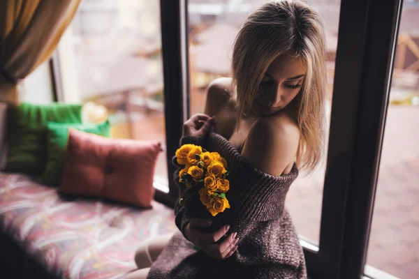 Retrato de jovem mulher bonita em pulôver cinza sentado na soleira da janela com flores amarelas — Fotografia de Stock