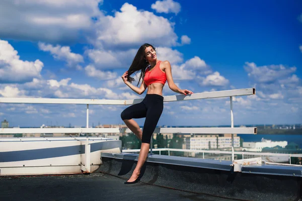 画像的金发坚强的女人，在做伸展运动以上大城市的摩天大楼的屋顶上，在清晨的运动服。女性运动和性感肌肉发达的身体。健身概念. — 图库照片