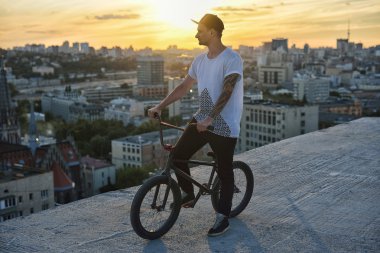 Genç yakışıklı hipster adam çatıda oturan ve günbatımı üzerinde bisiklet üzerinde poz