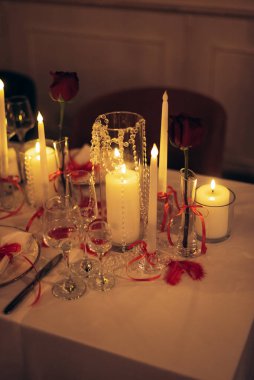 Tabaklar, kırmızı kurdeleler, cam tutacaklardaki mumlar, vazodaki tek güller ve romantik bir ortam yaratan kristal boncuklu zarif restoran ziyafeti masası..