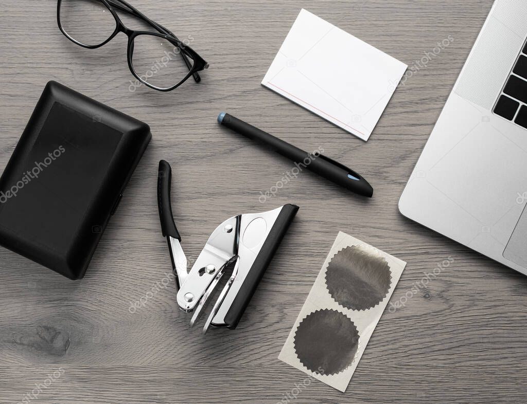 Flat lay of embossing hand tool with foil sticker, laptop, glasses, pen, and power bank on wooden table. Business and branding concept.