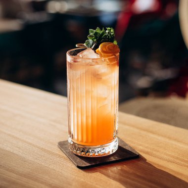 Portakallı tropikal kokteyli buz, nane yaprakları ve ahşap masada portakal dilimleri ile tazeliyorum. Ilık gün batımı canlı yaz içeceği atmosferi yaratır..