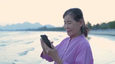 Tatil konsepti 4k Çözünürlük. Sahilde cep telefonuyla oynayan yaşlı bir Asyalı kadın..