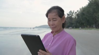Tatil konsepti 4k Çözünürlük. Yaşlı bir Asyalı kadın sahilde tabletle mutlu bir şekilde oynuyor..
