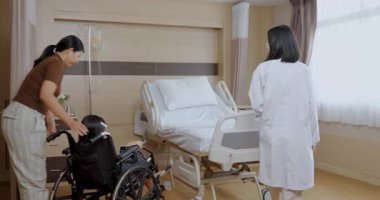 Hastane konsepti 4k Kararlılık. Asyalı bir anne, doktor muayenesinden sonra çocuğunu tekerlekli sandalyeyle hasta odasına getiriyor..