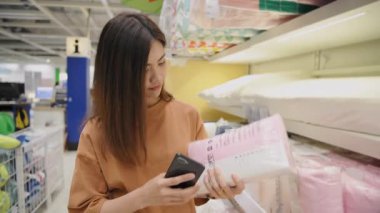 4k Kararlılığın alışveriş kavramları. Asyalı bir kadın alışveriş merkezindeki battaniyeleri karşılaştırırken cep telefonundaki fiyatları kontrol ediyor..