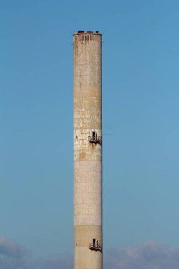 Çok yüksek harap olmuş, güçlendirilmiş beton baca. Mavi gökyüzü arka planındaki eski endüstriyel tesiste bulutların üzerinde yükselen iki açıklık ve güvenlik platformu var.