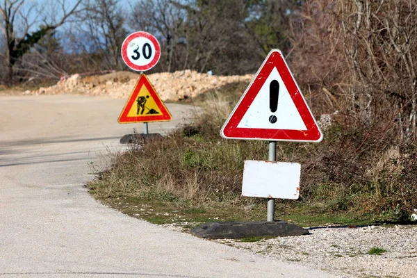 Dilapidated heavily used warning and road under construction with 30 speed limit road signs mounted on metal poles next to paved road surrounded with dry trees and other vegetation on clear blue sky background