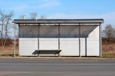 Parlak metal çerçeve ve kirli beyaz arka planla desteklenen modern otobüs durağının önü kuru çimenler ve açık mavi gökyüzü ile çevrili asfaltlı cadde soğuk kış gününde
