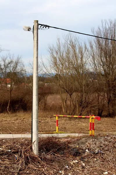 Kapalı kırmızı ve sarı şeritli rampanın yanında eski sokak lambası olan beton elektrik direği, arka planda kesilen dallar ve ağaçlarla çevrili yerel ormanın dar kaldırımlı girişini kapatıyor.