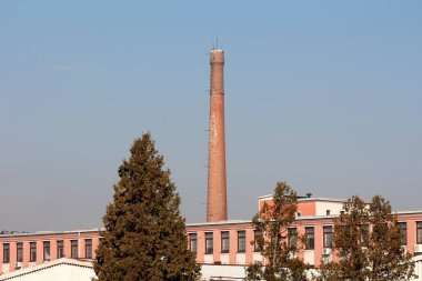 Kırmızı tuğlalar, uzun endüstriyel bacalar. Metal merdivenler yan tarafa monte edilmiş. Uzun, eski fabrika binası ve sıcak kış gününde ağaçların üzerinde yükseliyor.