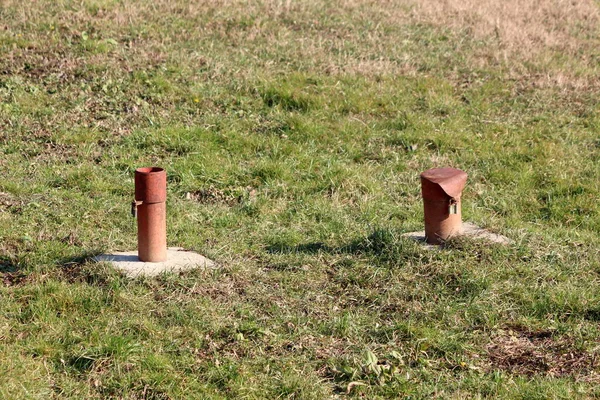 Two old rusted industrial metal pipes with broken padlock locked caps on top on hard concrete foundation surrounded with partially dry uncut grass on warm sunny winter day