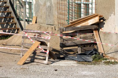 Kum yığını ve ahşap paletler ahşap tahtalar ve diğer inşaat malzemeleriyle harmanlanmış eski harap binanın yanında çakıl kaplı ön cephe ve paslı metal dış merdivenlerle çevrili.