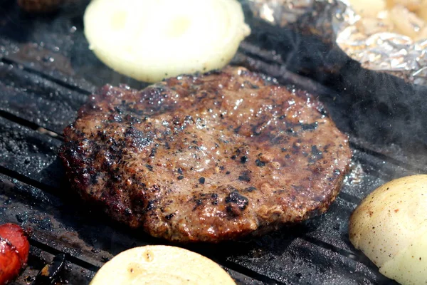 Homemade grilled hamburger patty covered with sauce and smoke next to onion rings and sausages grilling on top of outside charcoal barbeque grill on warm sunny spring day