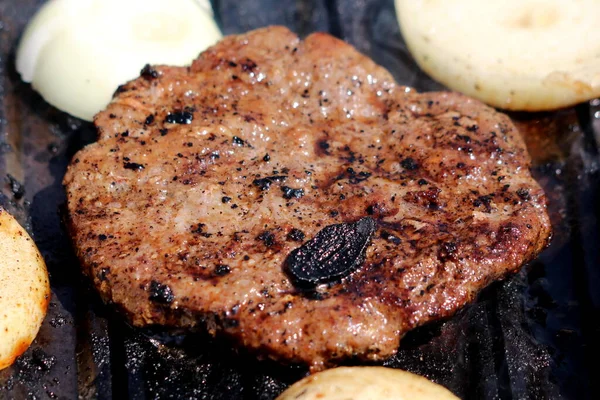 Grilled hamburger patty covered with sauce and smoke surrounded with onion rings grilling on top of outdoor charcoal barbeque grill on warm sunny spring day