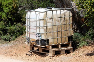 Ağır bir şekilde kullanılan eski beyaz orta büyüklükteki plastik kap ya da bükülmüş metal kafesli IBC plastik tankı yerel inşaat alanındaki ahşap paletlerin üzerine konmuş ve etrafı ağaçlar ve sürüngen bitkileriyle kaplı taşlarla çevrili.