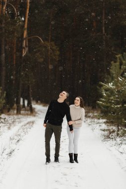 Gülücükler saçan genç bir çift, kış ormanında mesajlarınız için yer ararken karda yürüyorlar. Aşk, ilişki, kış tatili. Kış çifti fotoğraf fikirleri