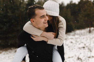Çam ormanının arka planında gülümseyen kadınını sırtında tutan mutlu bir adam. Aşk, ilişki, kış tatili. Kış çifti fotoğraf fikirleri