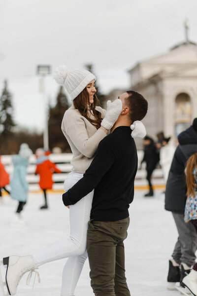 Мужчина держит улыбающуюся женщину на публичном катке. Женщина в белой вязаной шляпе и перчатках обнимает своего любовника. Любовная история. Зимнее веселье