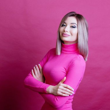Pembe uzun kollu bir kadın portresi pembe arka planda çapraz kollarla dikiliyor. Pembe arka planda bir kadın. Yüksek kalite fotoğraf
