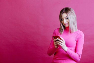 Pembe arka planda duran ve akıllı telefonuna bakan bir kadın. Mesajın için pembe arka planda bir kadın. Yüksek kalite fotoğraf