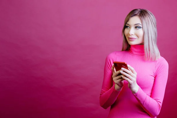 Woman in the pink longsleeves standing on the pink background with phone in her hands and looking to the side. Woman over pink background with place for your text. High quality photo