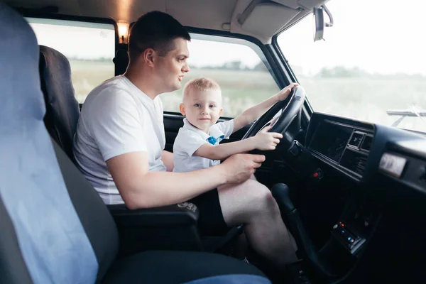Father driving a car with his little son. Little boy sitting on the fathers legs and turning the steering wheel in the car. Happy father and son