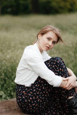  Genç ve şık bir kız arka planda bir ağaç kütüğünde oturur ve kameraya bakar. Yakın plan fotoğraf. Metnin için yer.