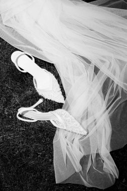Bir çift güzel beyaz gelinlik ayakkabısı yumuşak çimenlerin üzerinde dalgalı bir düğün peçesinin yanında duruyor, bir an için zarafet ve beklenti yakalıyor..