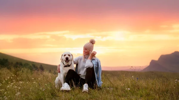 Bir kız ve bir labrador dağlarda gün batımının arka planında oturuyorlar.