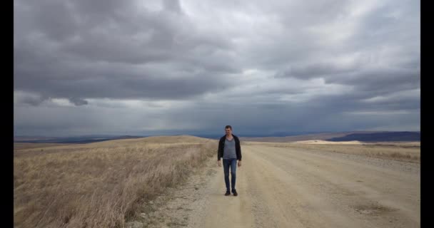 Un tipo camina a lo largo de un camino de campo en gafas un fuerte viento sopla nublado — Vídeos de Stock
